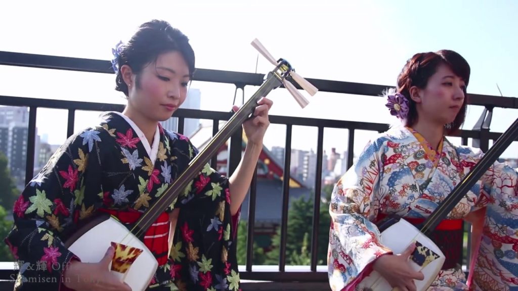 A photo of Ki&Ki, sitting on a balcony, holding their shamisens, mid-performance of the traditional song Tsugaru Jongara Bushi.