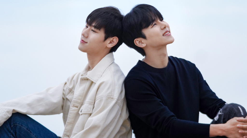 A promotional photo from the film Wish You of actors Lee Sang and Kang In Soo sitting back to back and smiling off into the distance.