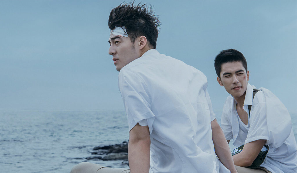 A photo still from Your Name Engraved Herein. Lead actors Edward Chen and Jing-Hua Tseng are sitting on a beach, with the ocean behind them, wearing white shirts and looking off into the distance.