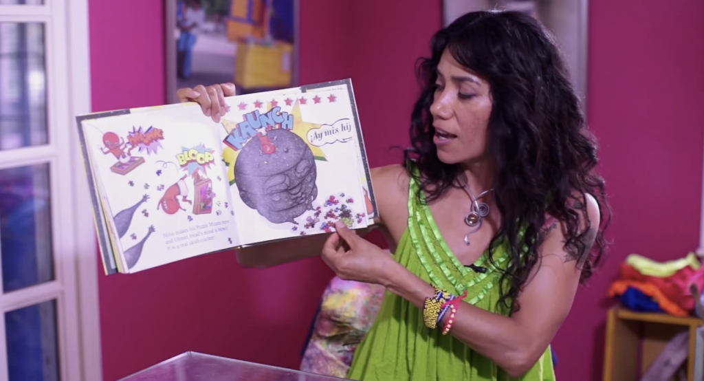 A photo of author Yuyi Morales holding up her children's book Niño Wrestles the World. She is looking at the open pages, and is mid-read.