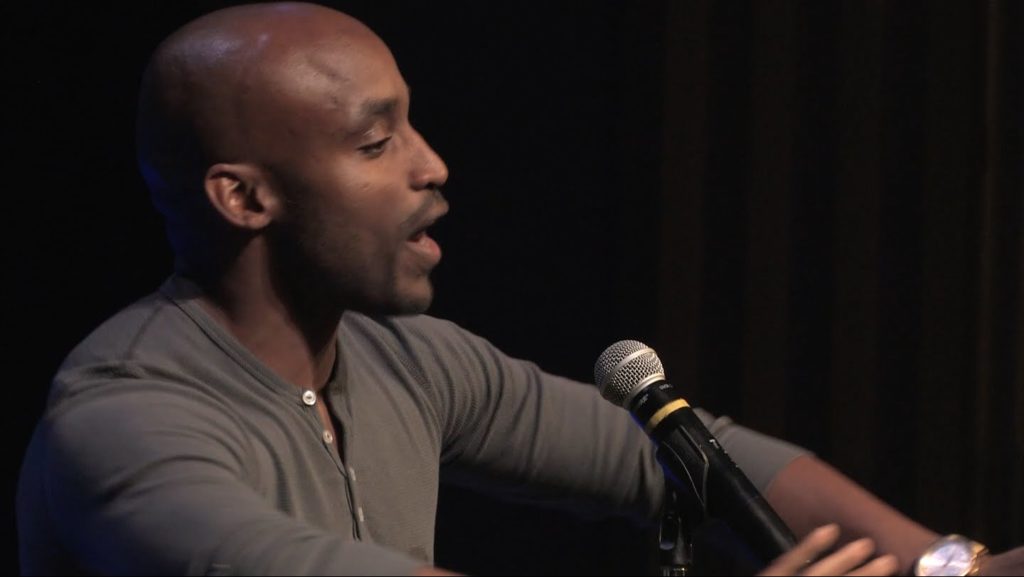 A photo of Javon Johnson, mid-performance, onstage, speaking into a microphone.