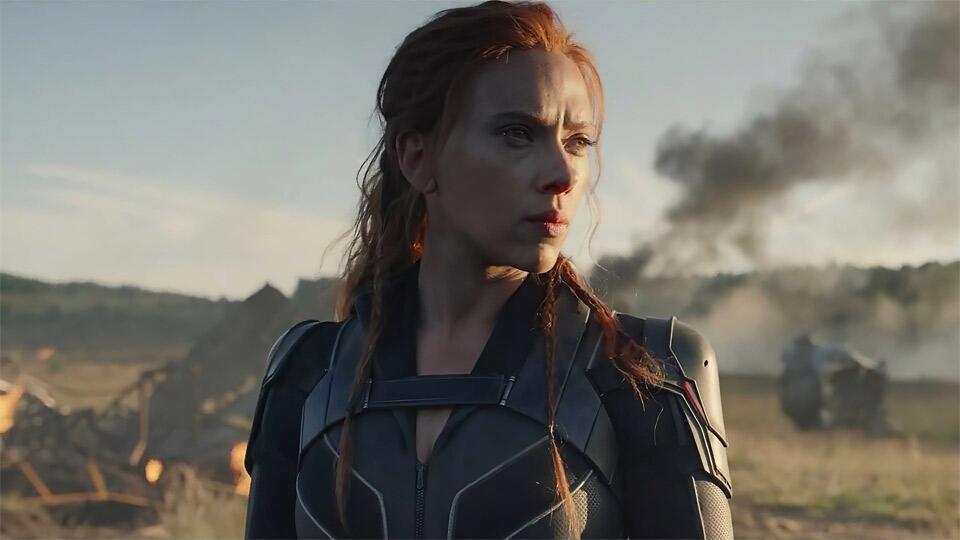 A photo still from the film Black Widow. the photo is of Scarlett Johansson in costume, standing in a field and staring off camera as the wreckage of a battle smokes behind her.