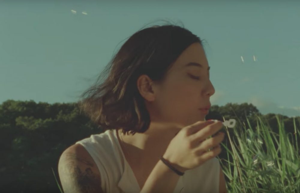 A photo still from the music video of The Body is a Blade by Japanese Breakfast. Lead singer Michelle Zauner is blowing bubbles in a field under a blue sky.