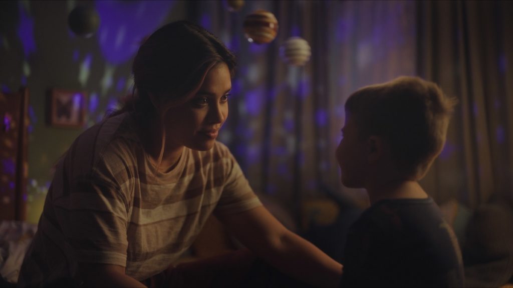 A photo still from Let's Be Tigers. Lead actors Otmara Marrero and Dash McCloud are sitting on the floor in a little boy's room, smiling at each other.
