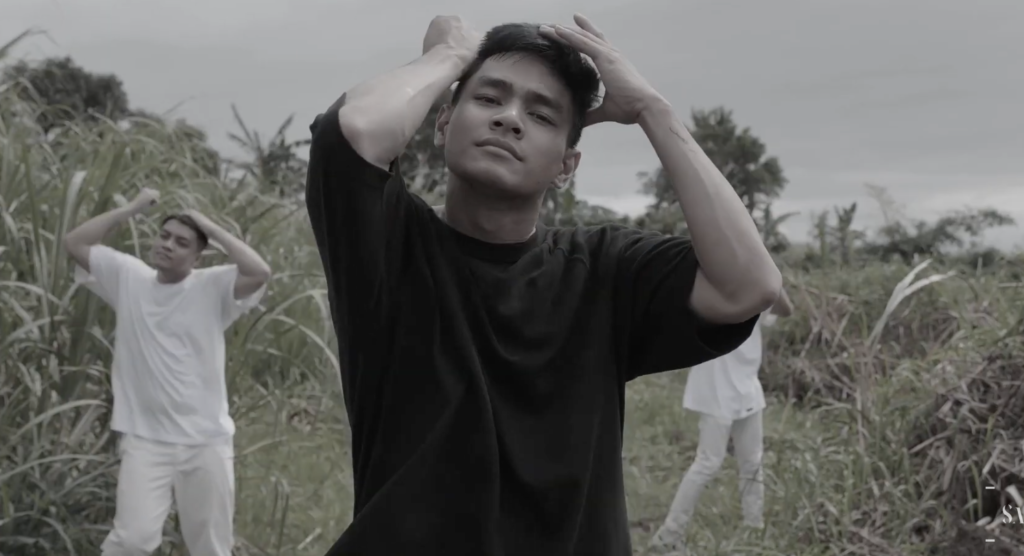 A photo of dancer Shim Abrio, mid-performance, standing in the middle of a field with tall grass. He's wearing all black, and two back up dancers behind him are wearing all white.