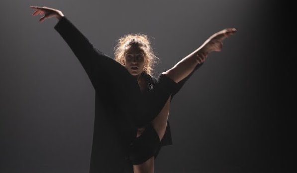 A photo of dancer Tess Voelker mid-performance, holding one leg up in the air to the side of her body.