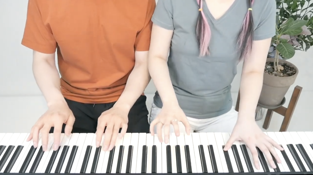 A close up photo of Bella & Lucas as they play the piano together. They are only seen from the neck down as they sit at the piano.