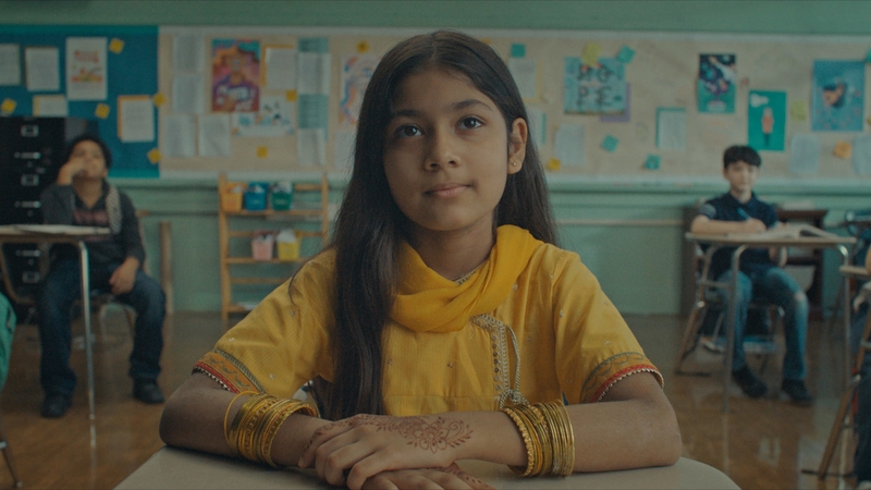 A photo still from American Eid. Lead actress Shanessa Khawaja is sitting at a desk in a classroom, hands folded in front of her, looking straight ahead just above the camera.
