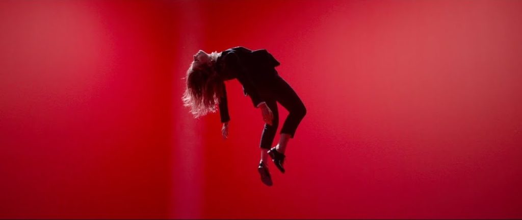 A photo still from the video for Saint Claude by Christine and the Queens. A woman in a black suit is suspended mid-air, back arched, against a red background.