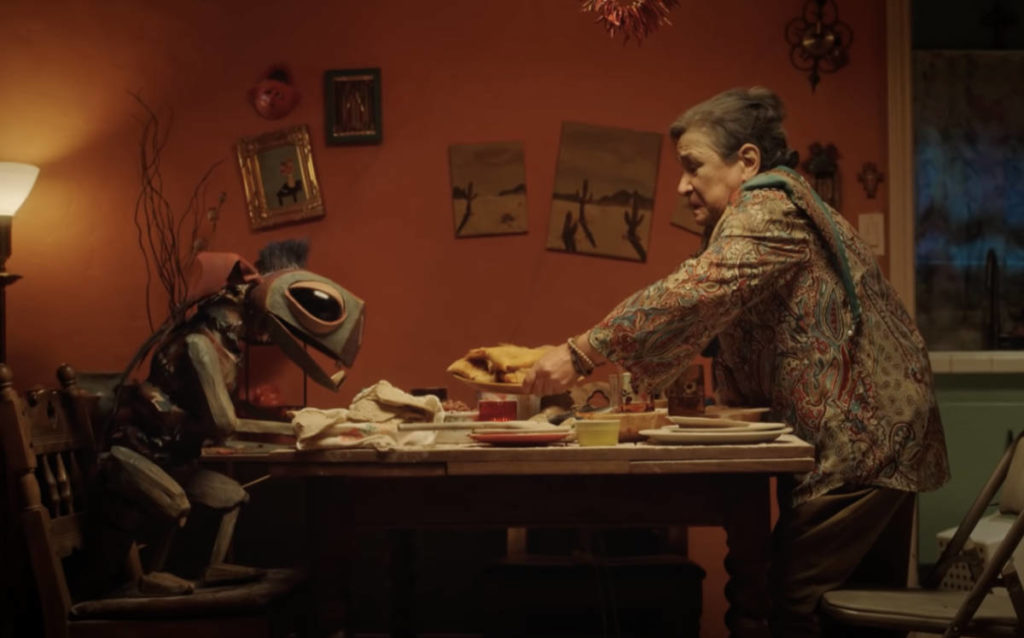A photo still from the short film The Last of the Chupacabras, showing the chupacabra and the lead actress sitting at a table having a meal together.