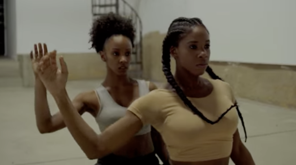 A photo still from Devoted, a dance film. Dancers Annellyse Munroe and Aqura Lacey are kneeling on the ground, one behind the other, with their right hands raised.