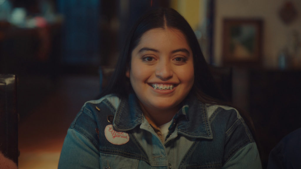 A photo still from the short film Growing Fangs of lead actor Keyla Monterroso Mejia. It is a close up photo of her sitting at a table in a denim jacket, smiling broadly.