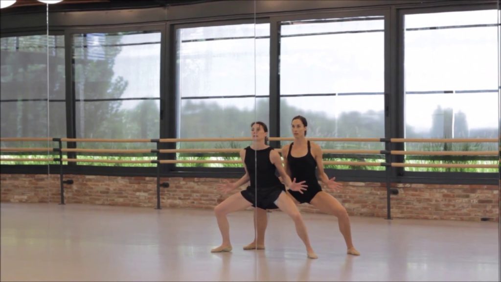 A photo still from the video Back Forward Back, choreographed by Juliano Nunes. The photo shows two female dancers in plain black outfits, dancing in a studio. The camera is filming them through the floor to ceiling mirrors in the dance studio.