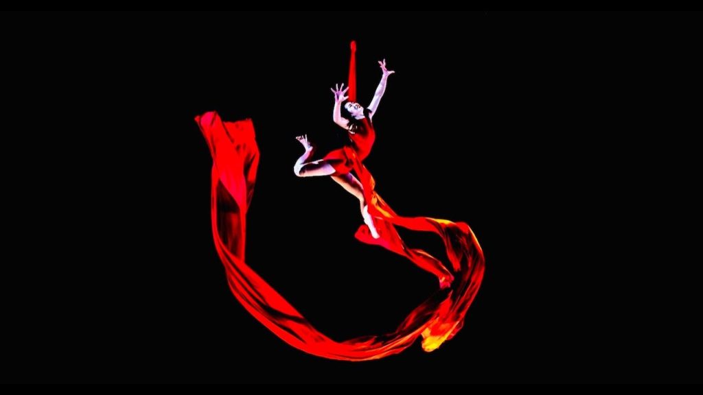 A photo of aerial silks performer Mizuki Shinagawa, suspended in the air by red silks, the material flowing around her, against a black backdrop.