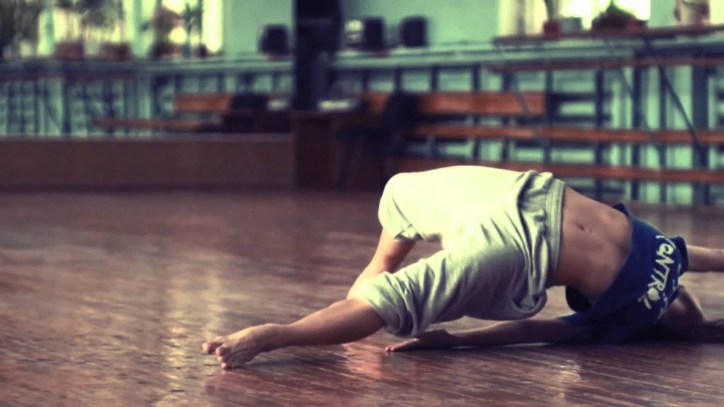 A photo still from the video of Olga Kuraeva doing an improvised contemporary ballet dance. In the photo, she is on the ground in a dance studio, her back arched to raise her body in the air.