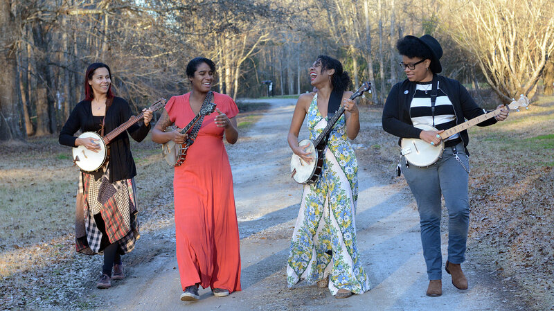 A photo of the four members of Our Native Daughters. They are standing on a dirt path in a park, laughing and holding their instruments.