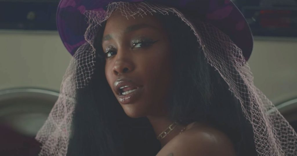A photo still from the music video for Drew Barrymore by SZA. The photo is a close up of SZA looking over her shoulder straight into the camera. She is wearing a purple hat with a veil.