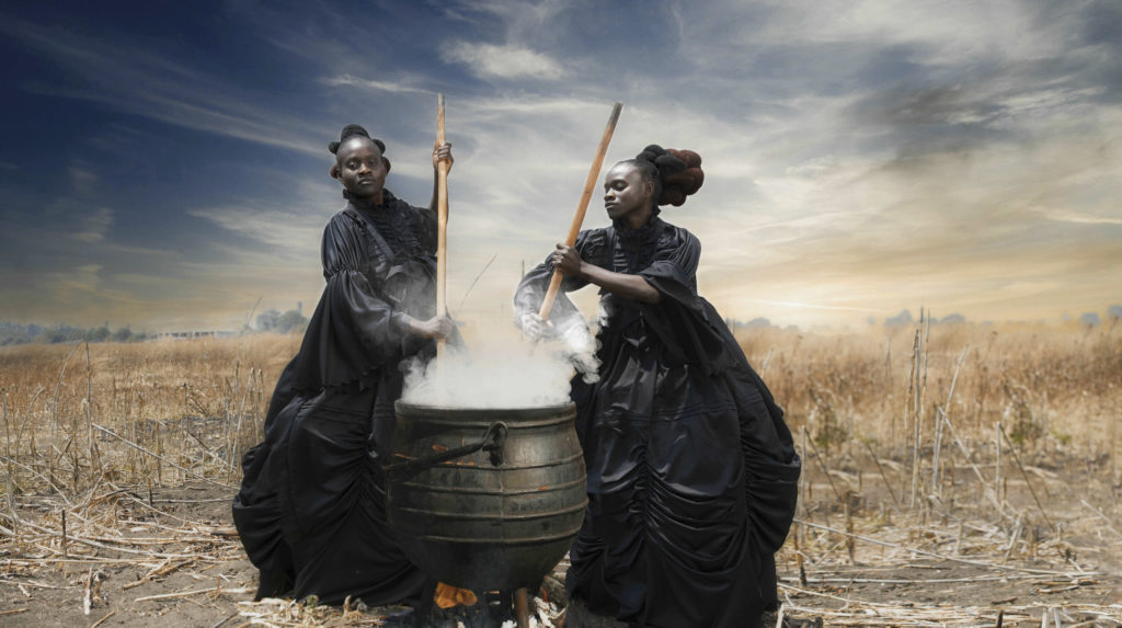 The photo That Evening Sun Goes Down by Tamary Kudita. The photo is of two Black women in black Victorian dresses, stirring a boiling cauldron.
