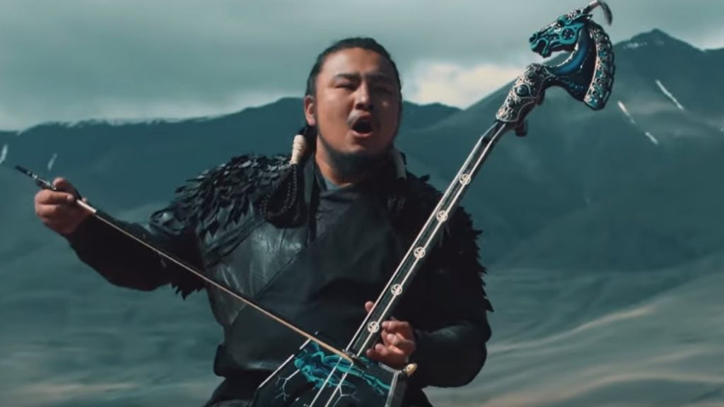 A photo still from the music video of Yuve Yuve Yu by The Hu. It shows a musician playing a morin khuur and singing among a backdrop of mountains.