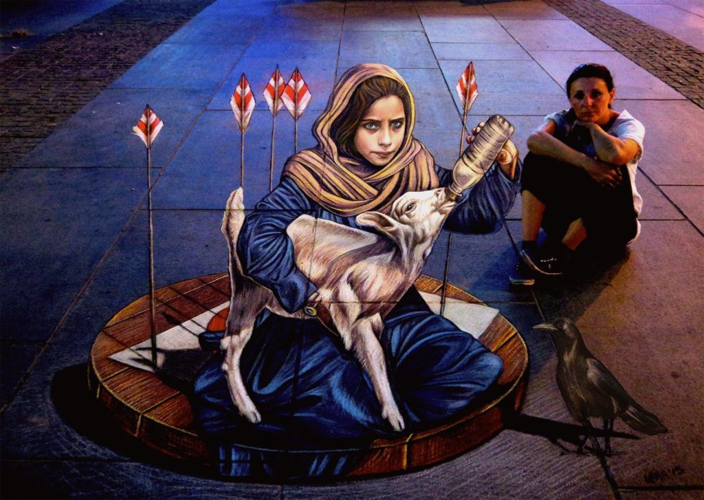 A photo of artist Vera Bugatti sitting on the ground next to an image she painted onto the pavement. The image is of a young girl feeding a baby goat with a bottle.