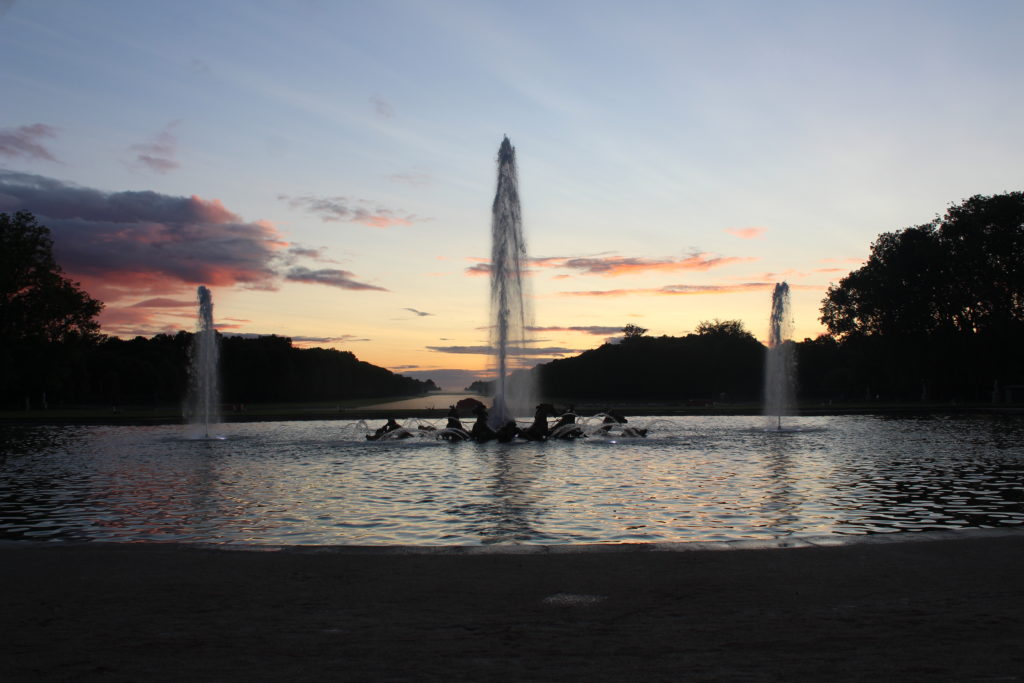 A photo of the fountains at the Versailles Fountain Show. Three jets of water and being sprayed into the air, as the sun sets in the background.