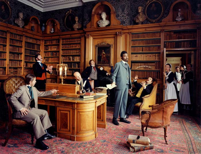 A photo from the series Diary of a Victorian Dandy by Yinka Shonibare. The photograph shows a group of men in Victorian dress meeting in a library while several maids peek in through the door. Yinka Shonibare is standing in the centre of the photograph.