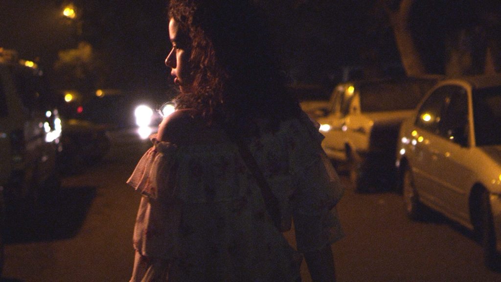 A photo still from Youth. The lead actress is shown from behind, walking down a city street with cars parked on either side. It is night, and her face is illuminated by the head lights of an approaching car.