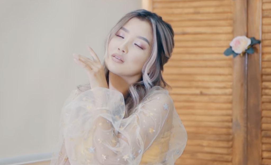 A photo still from the music video Life Goes on by Aidana Deka. It is a close up of Aidana Deka, wearing a pale pink dress, holding a hand up to her face and closing her eyes.
