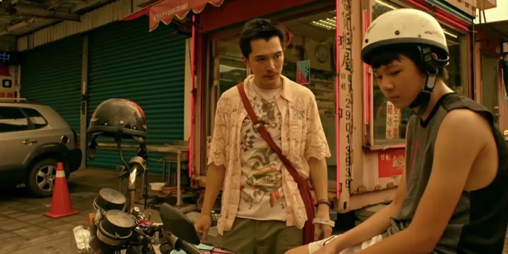 A photo still from the film Dear Ex. Actor Joseph Huang is sitting on a motorcycle, wearing a helmet and looking down and away from Roy Chiu who is standing next to the motorcycle and looking intently at him.