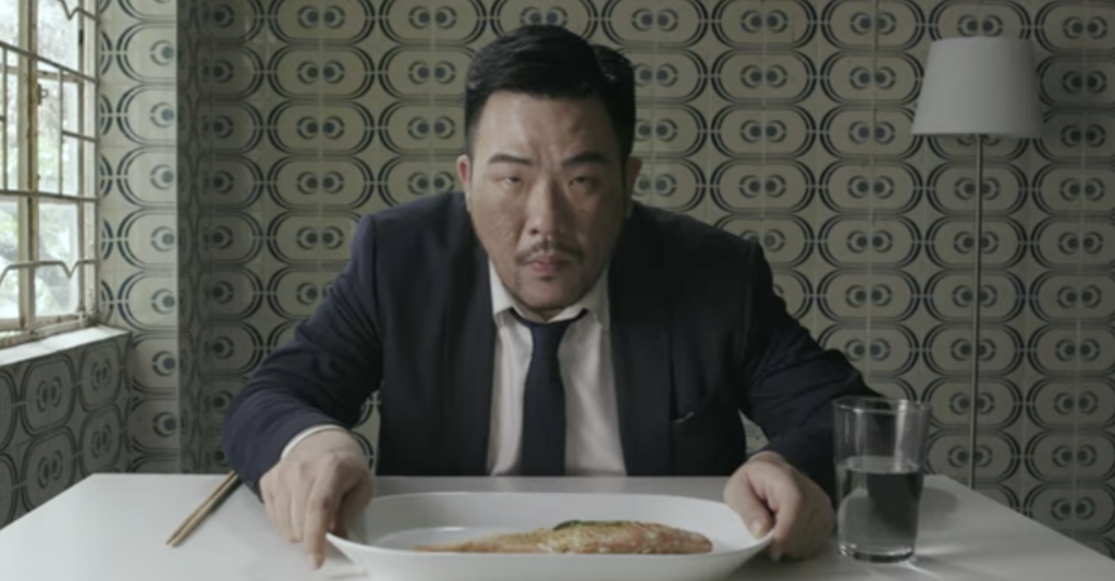 A photo still from the dance film Fish. A man is sitting at a table, a plate of food and a glass of water in front of him, and he's leaning over the food to stare straight into the camera.