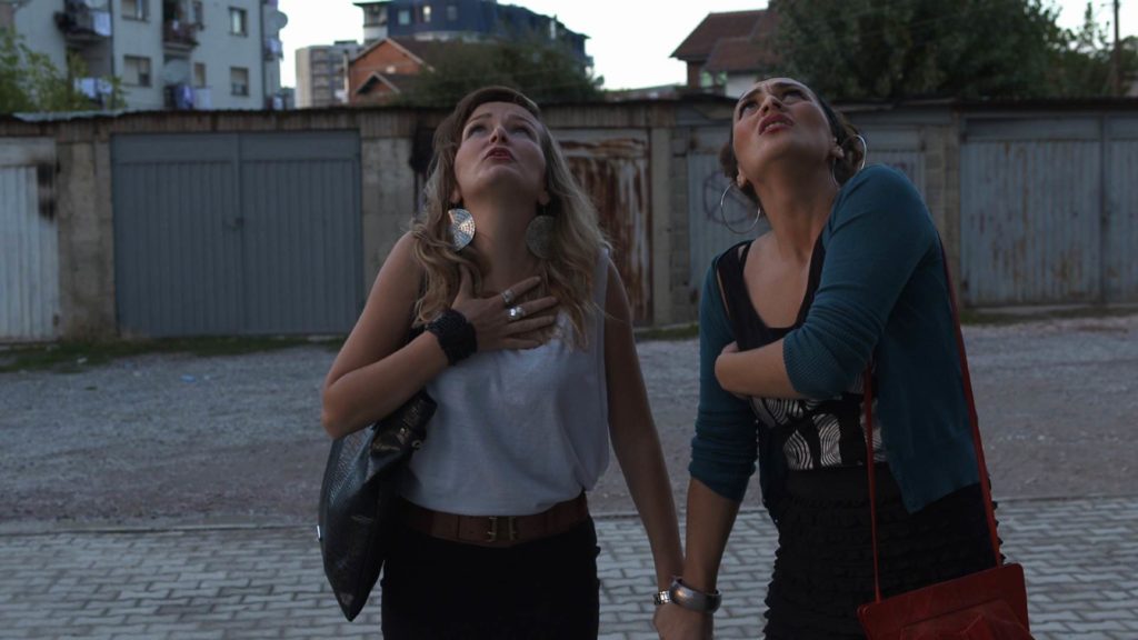 A photo still from the short film Balcony by Lendita Zeqiraj. Two women stand on a road, holding hands, and looking up at a building in horror.