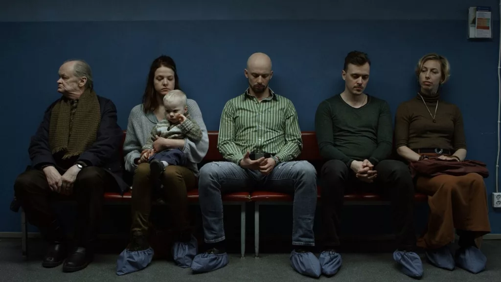 A photo still from the short film True by Marta Pulk. It shows five people sitting on a bench in a hallways. Their backs are all to a blue wall. There are three men and two women, one of whom is holding a baby.