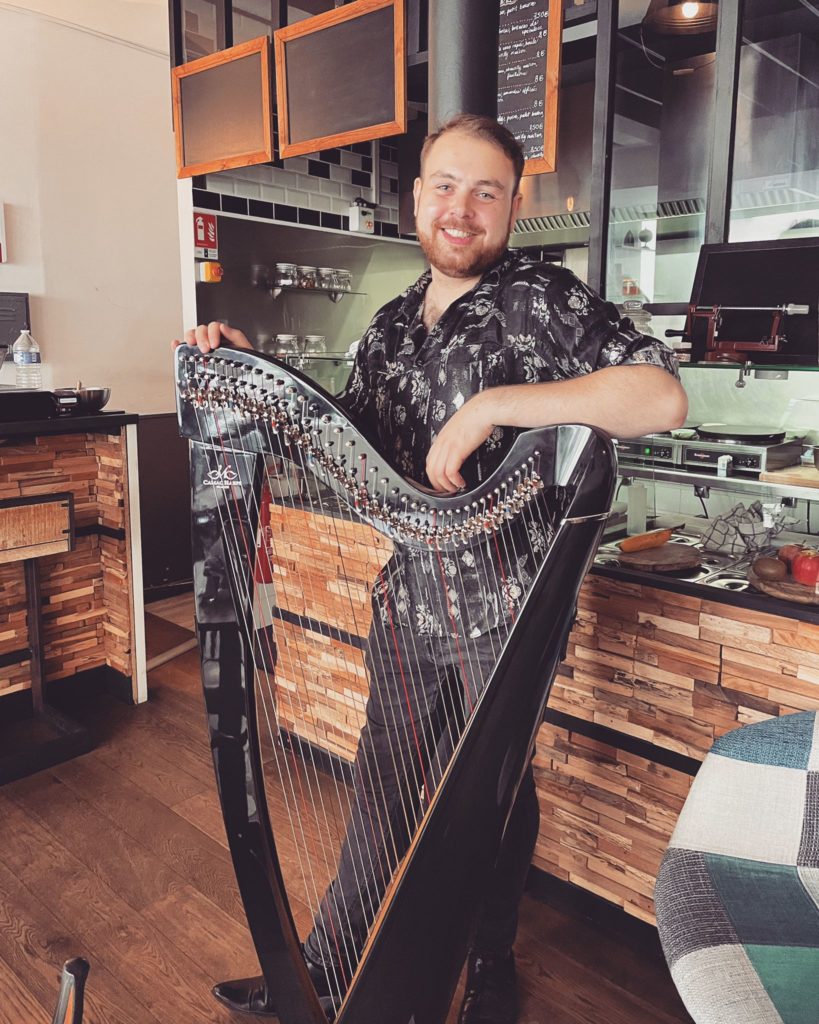 A photo of the musician Stiof, arm casually resting on his harp, standing in a bar and smiling directly into the camera.