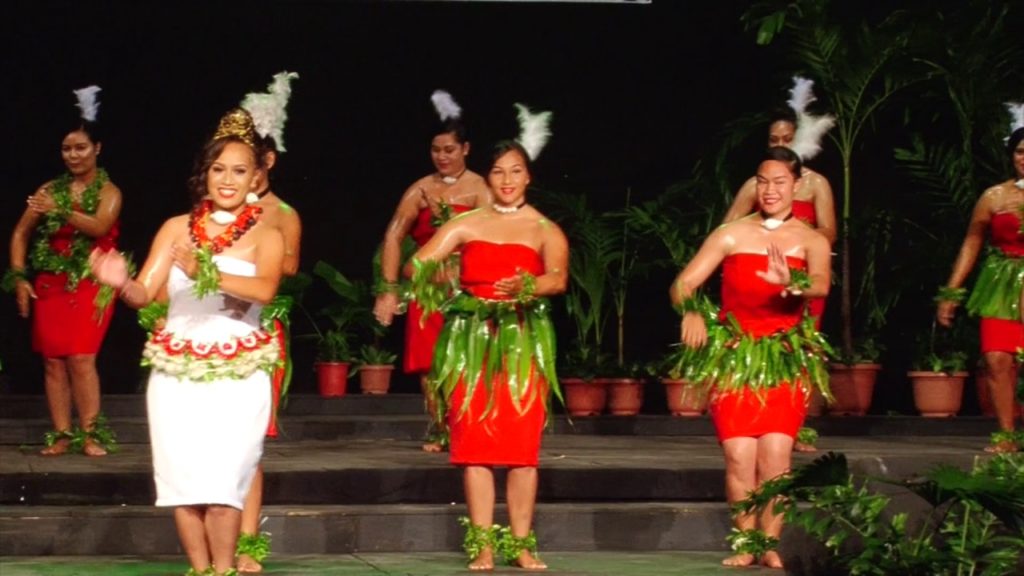 A photo of several female dancers performing Tau'olunga. They are all wearing red dresses with green grass skirts, with the exception of one on the left who is wearing a white dress.