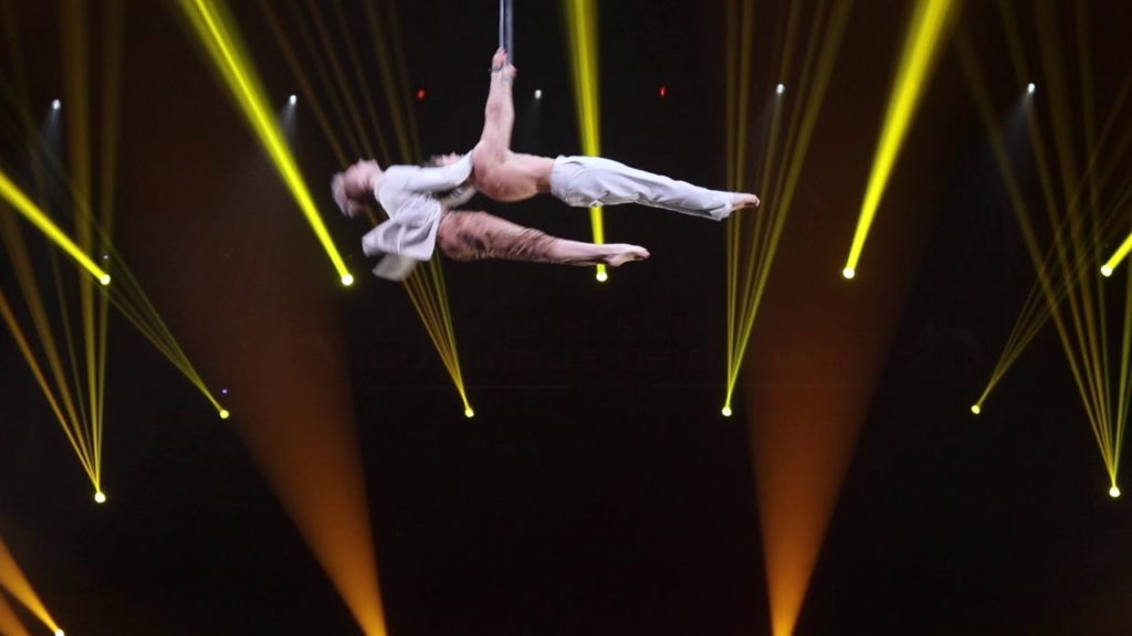 A photo still from the performance of The Same by an aerial straps duo at the 2019 Festival Mondial du Cirque De Demain in Paris. The two performers are suspended in the air by two straps, their bodies perfectly vertical, with one only holding onto the other's shoulders.