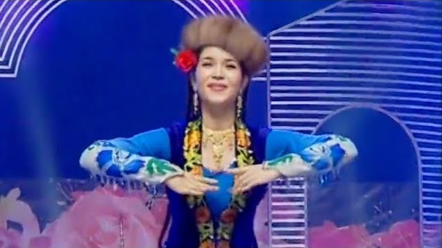 A photo of a Uyghur woman, mid-performance, wearing traditional Uyghur clothing, mostly in blue, and a fur hat with a rose tucked behind one ear.