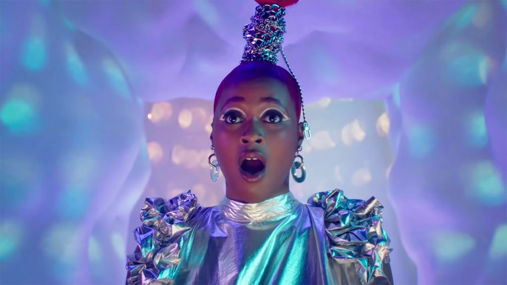 A photo still from the music video Link by Tierra Whack. The photo is a close up photo of Tierra Whack, dressed in a silver costume, staring off camera open mouthed.