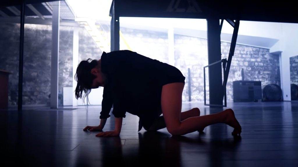 A photo still from the dance video by Marie Bugnon. She is on her hands and knees on the floor in a studio, mid-performance.