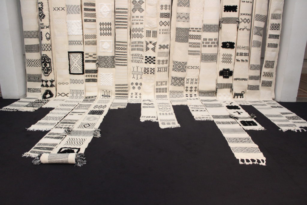 A photo of an artwork made of strips of white fabric with black detailing and symbols on them. When the strips reach the ground, they extend outward on the floor in varying lengths.