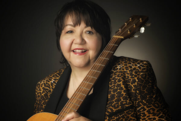 A close up photo of Norine Braun. She is smiling at the camera and holding a guitar.