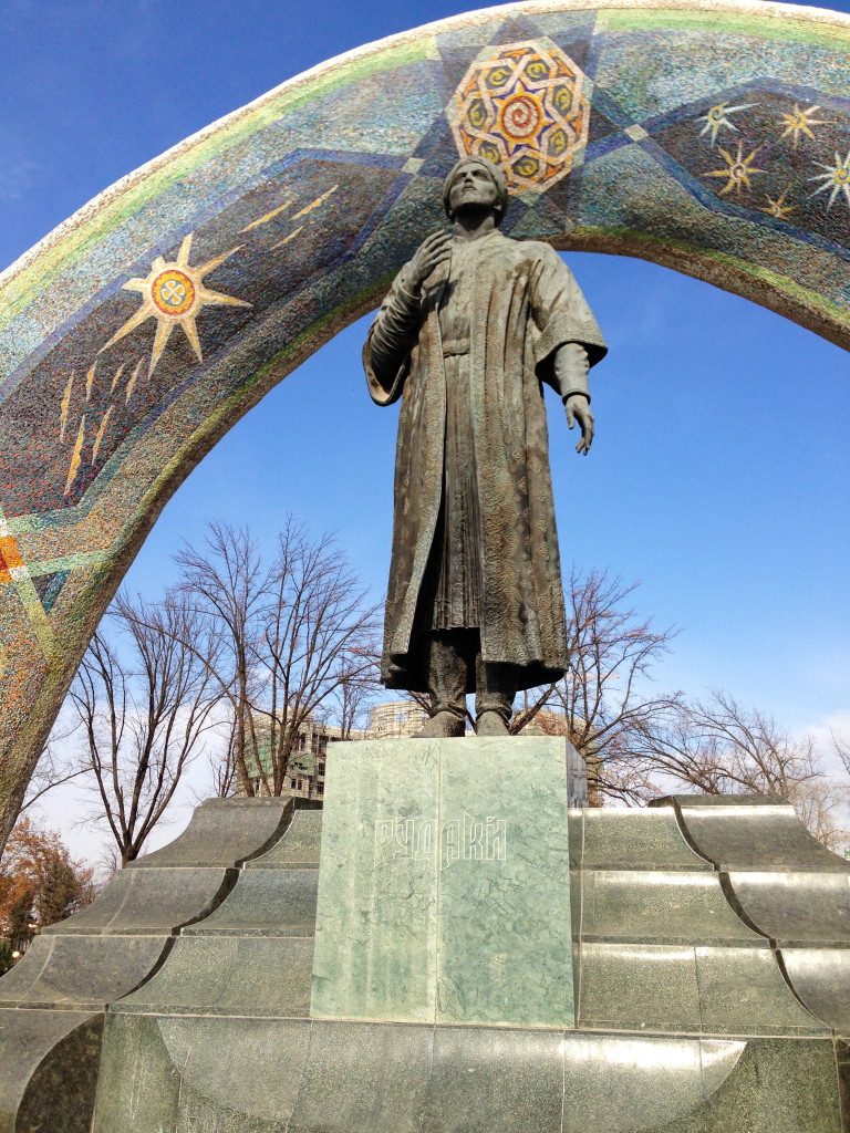 A photo of a statue of Rudaki, standing on a pedestal, underneath a decorated arch.