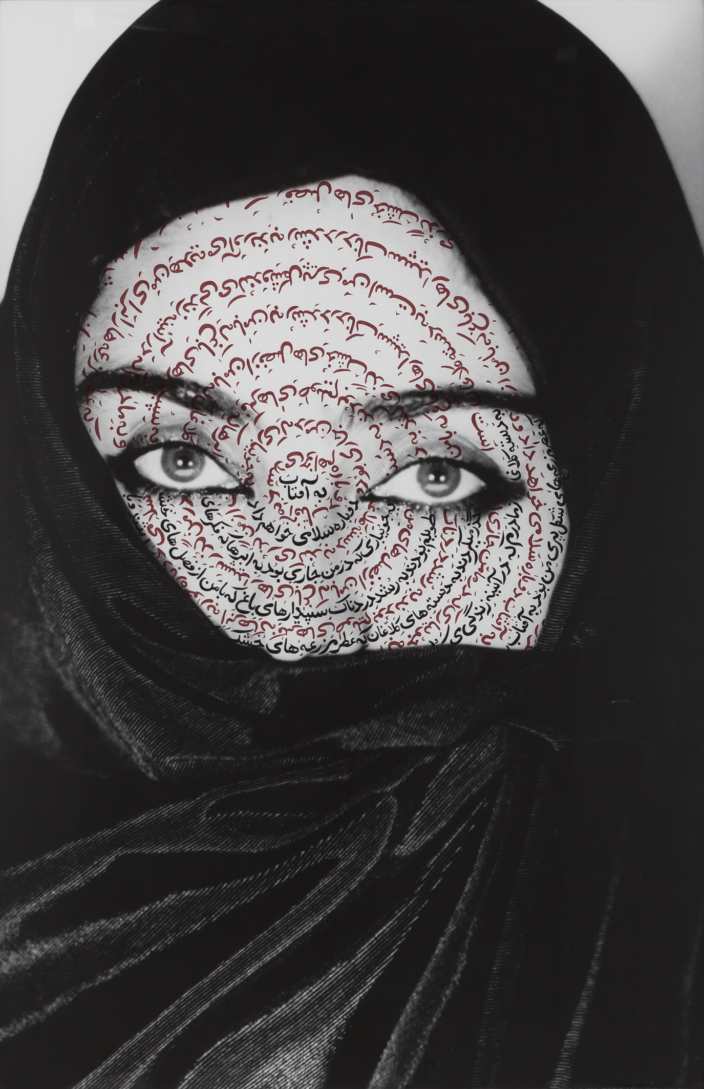 A photo by Shirin Neshat. It is a close up of a woman wearing a black headscarf. Her eyes are looking straight into the camera, and her face is covered in Arabic calligraphy.