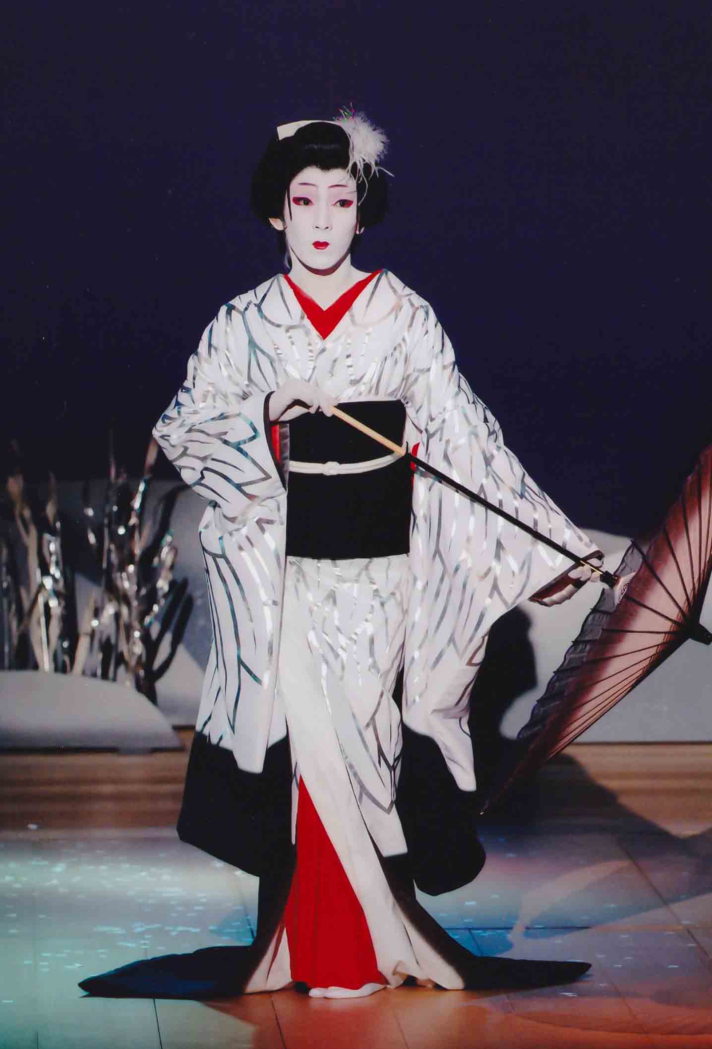 A photo of an onnagata performer. The actor is onstage in full dress, hair, and make up, holding an umbrella to the side.