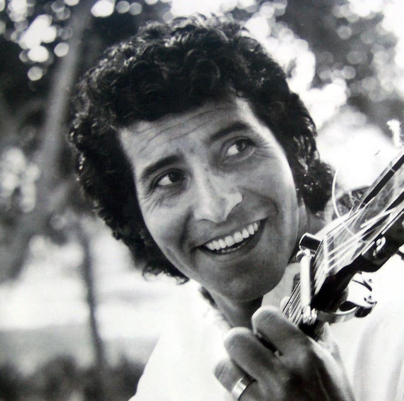 A close up black and white photo of Victor Jara sitting outside holding a guitar.