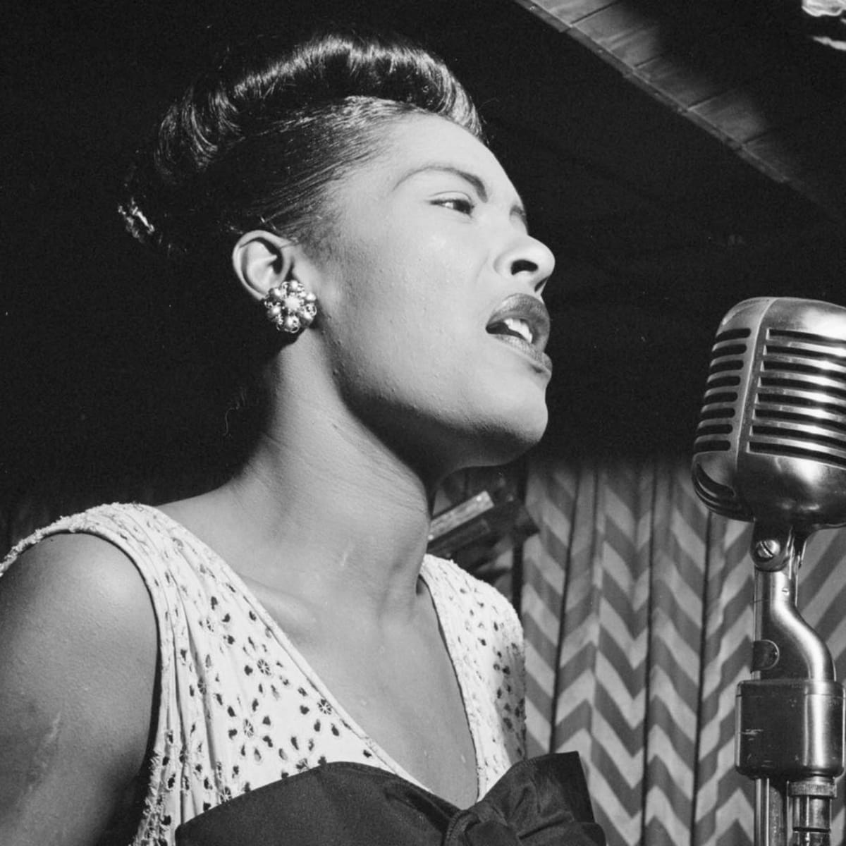 A black and white photo of Billie Holiday onstage, singing into a microphone.