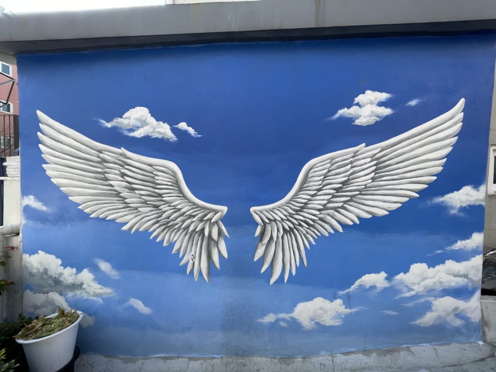 A photo of a mural in the Ihwa Mural Village. The mural shows a pair of white wings spread out against a blue sky background.