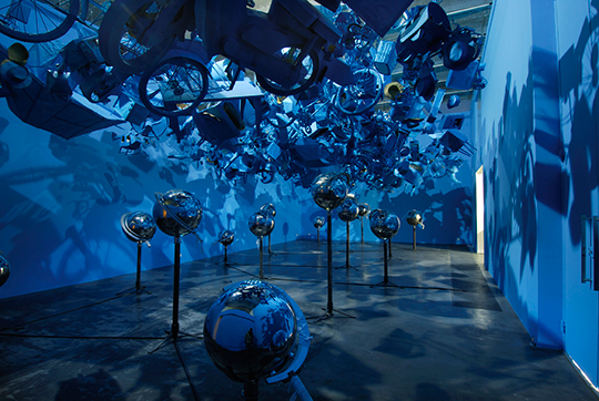 A photo of an art installation by Jun Nguyen-Hatsushiba. There are dozens of blue cycles suspended in the air in a room with blue walls.