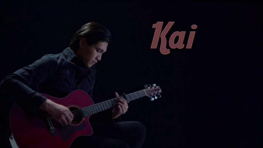 A photo of the musician Kai. He is sitting onstage, a black background behind him, playing a guitar.