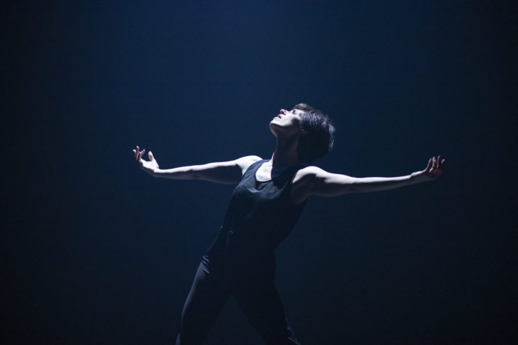 A photo of Marie-Claude Pietragalla onstage during a performance of La Femme Qui Danse. She is wearing all black, her arms are outstretched to the sides, and she is looking up.