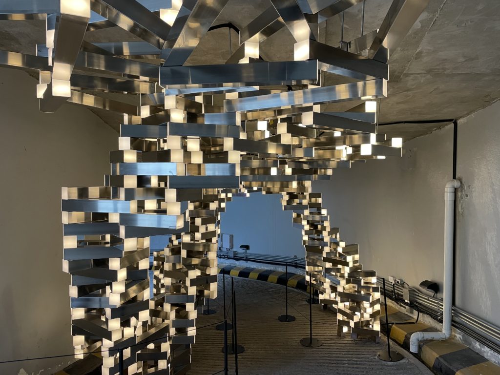 A photo of the artwork Net of Life - Arch by Joohyun Kim. The artwork is a large, floor to ceiling sculpture of metal pipes stacked on top of each other like legos.